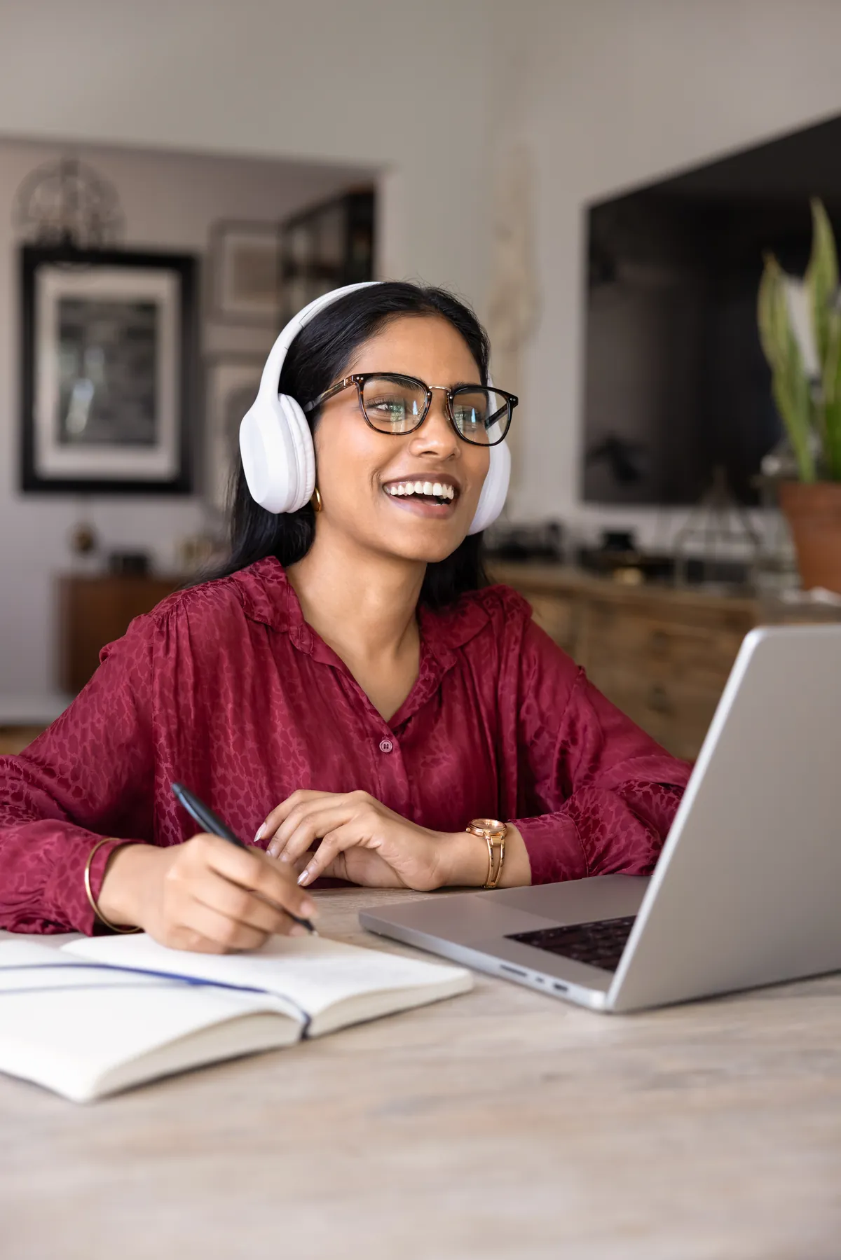 Woman wearing headphones and taking notes while using a laptop at home.